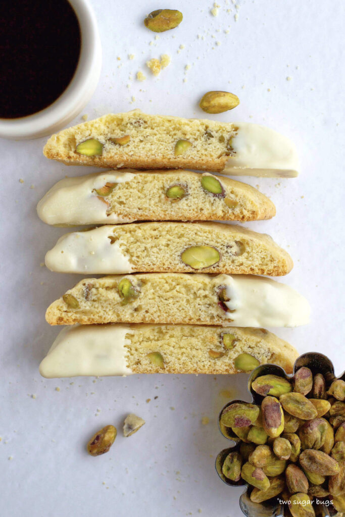 a stack of lemon pistachio biscotti with a cup of coffee and bowl of pistachio nuts