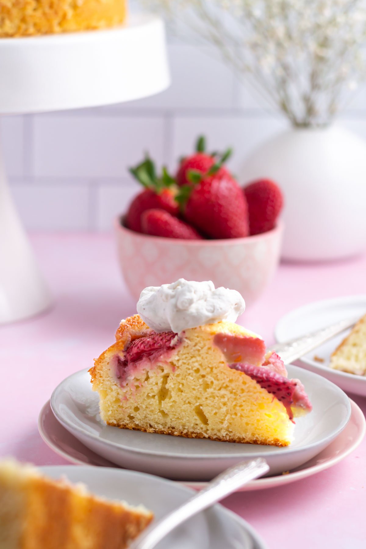 slice of strawberry vanilla cake with whipped cream on a plate with bowl of strawberries in the background