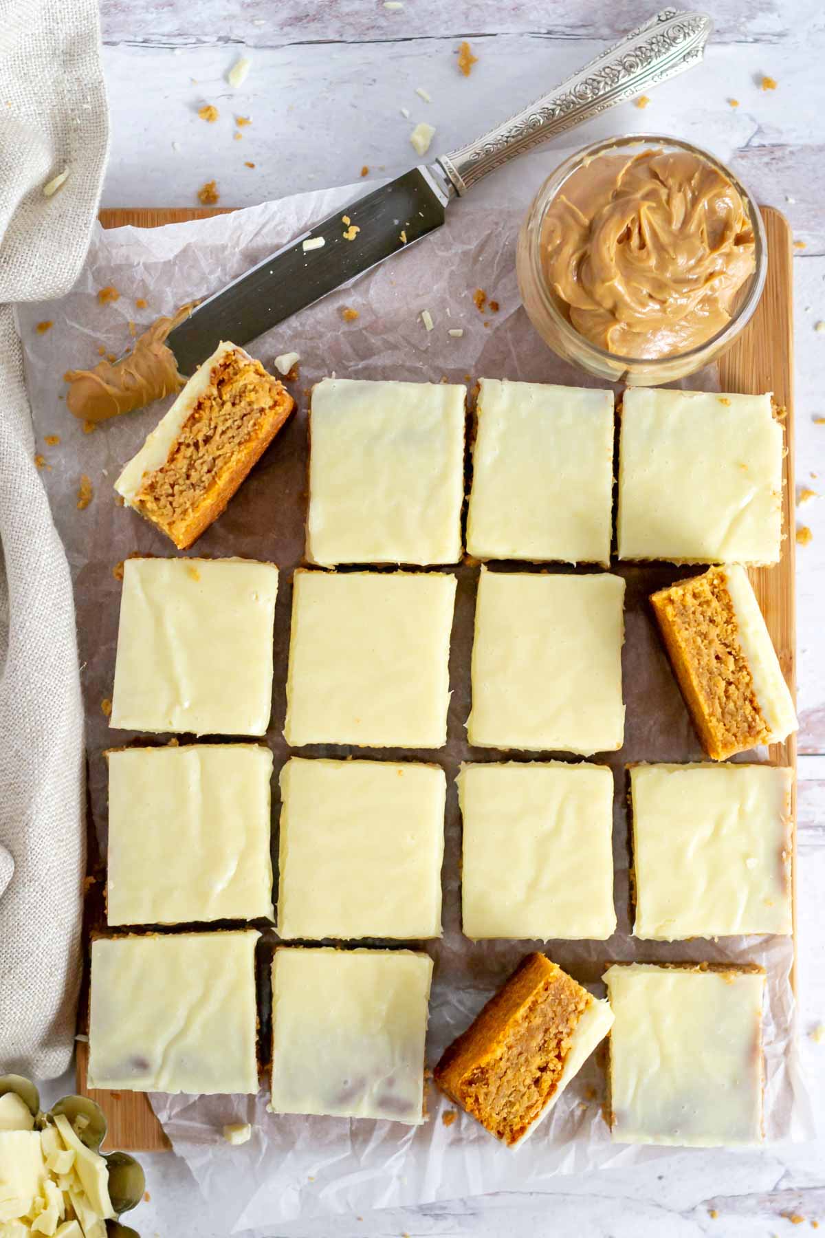 blondie bars on a cutting board with a knife, bowl of peanut butter and chopped white chocolate