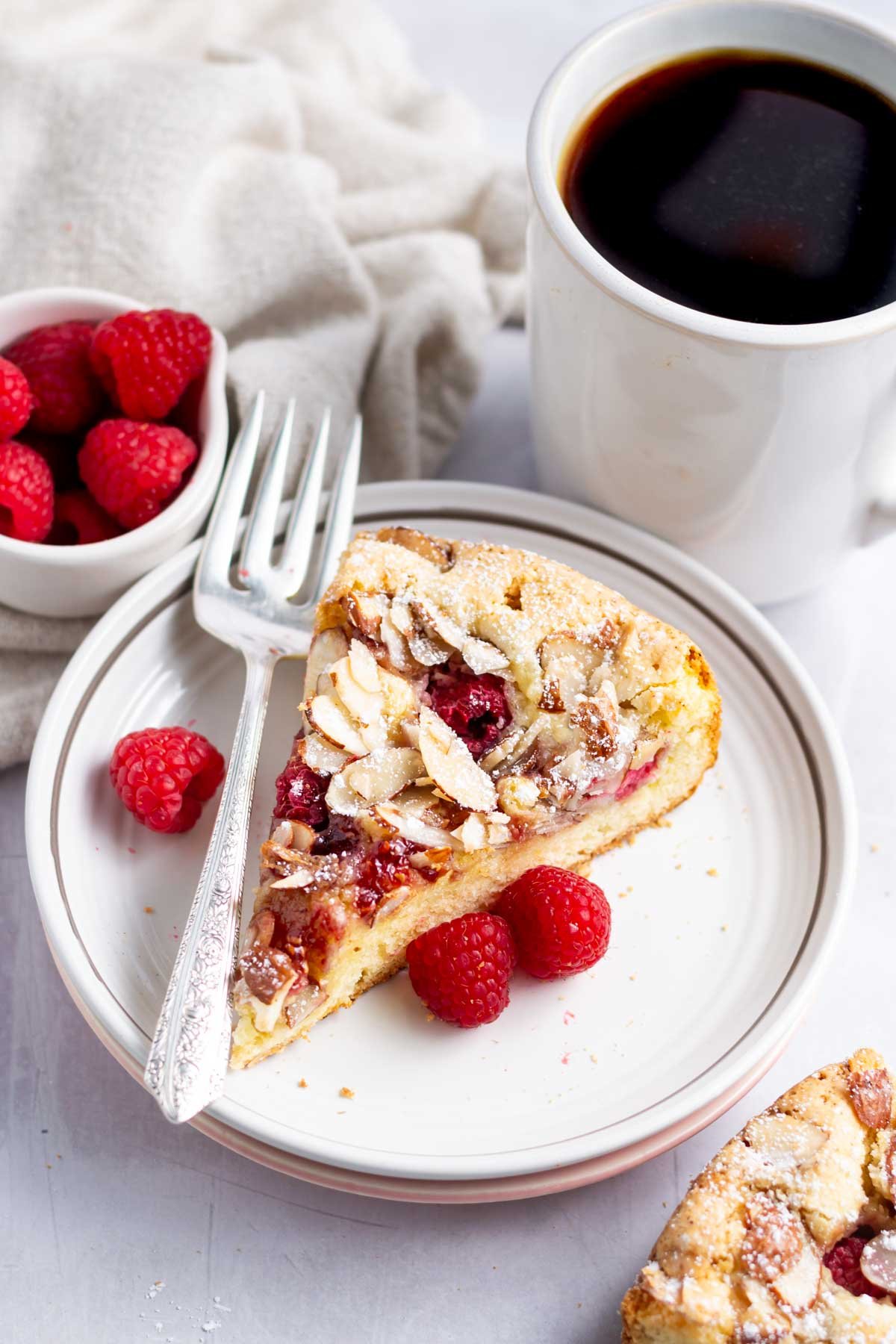 slice of raspberry almond cake on a plate with a fork and a cup of coffee
