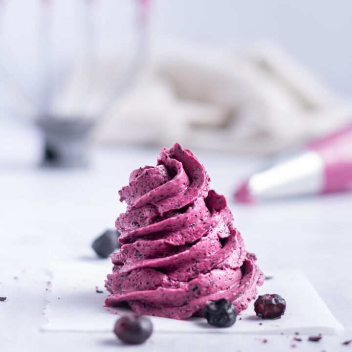 swirl of blueberry frosting on a piece of parchment paper with freeze dried berries and a piping bag in the background