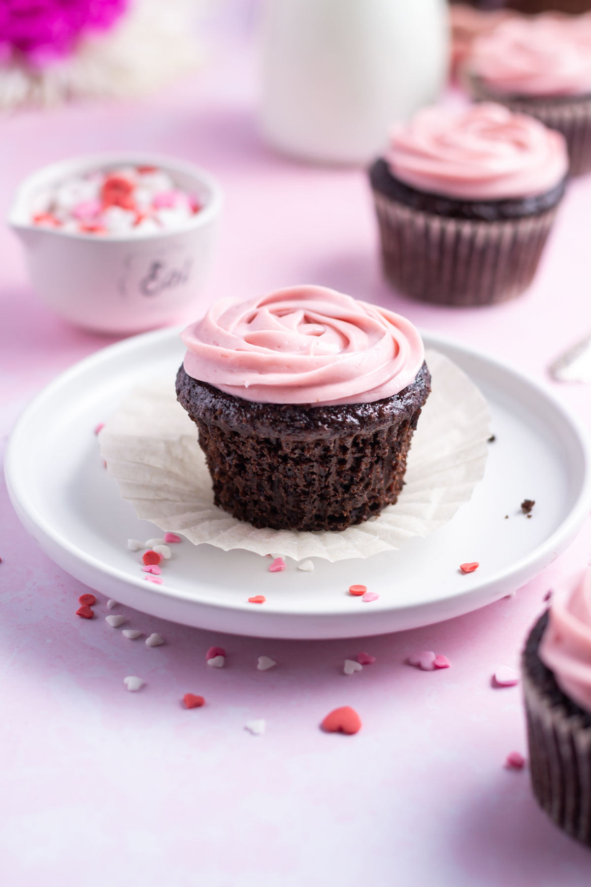 chocolate cupcake with the wrapper missing on a plate with a bowl of sprinkles and milk in the background