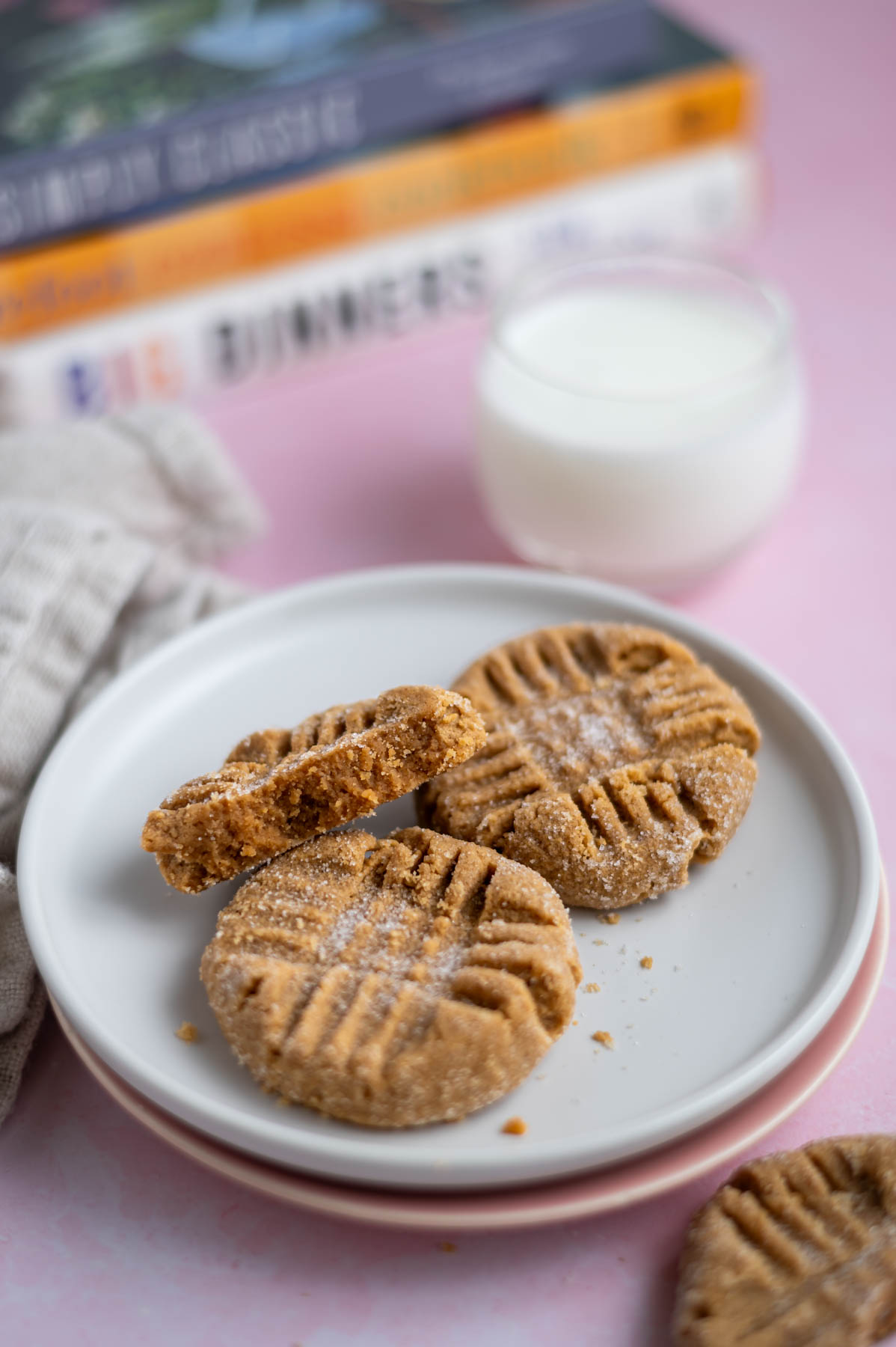 three peanut butter cookies on a plate with a glass of milk and cookbooks in the background