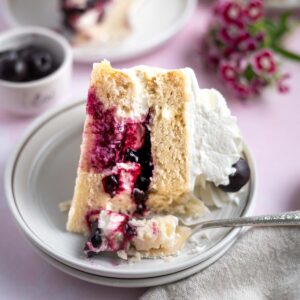 slice of white forest cake on a plate with a fork and flowers and a bowl of cherries in the background