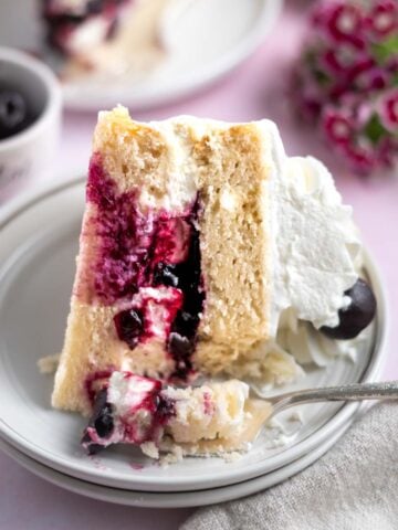 slice of white forest cake on a plate with a fork and flowers and a bowl of cherries in the background