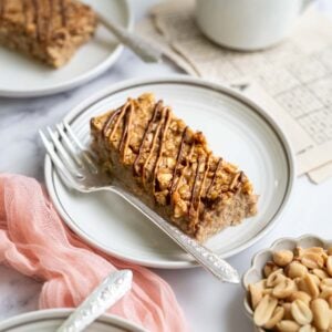 slice of baked peanut butter oatmeal on a plate with a fork. A bowl of peanuts and coffee sitting nearby.