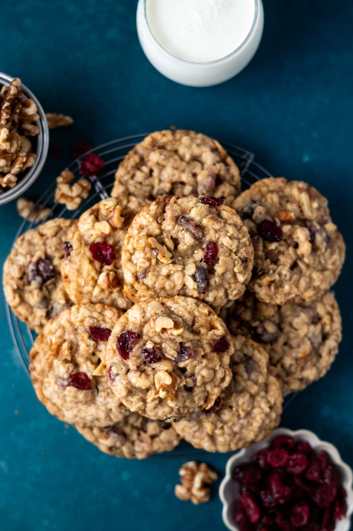 cranberry walnut oatmeal cookies on a cooling rack with a glass of milk and a bowl of dried cranberries