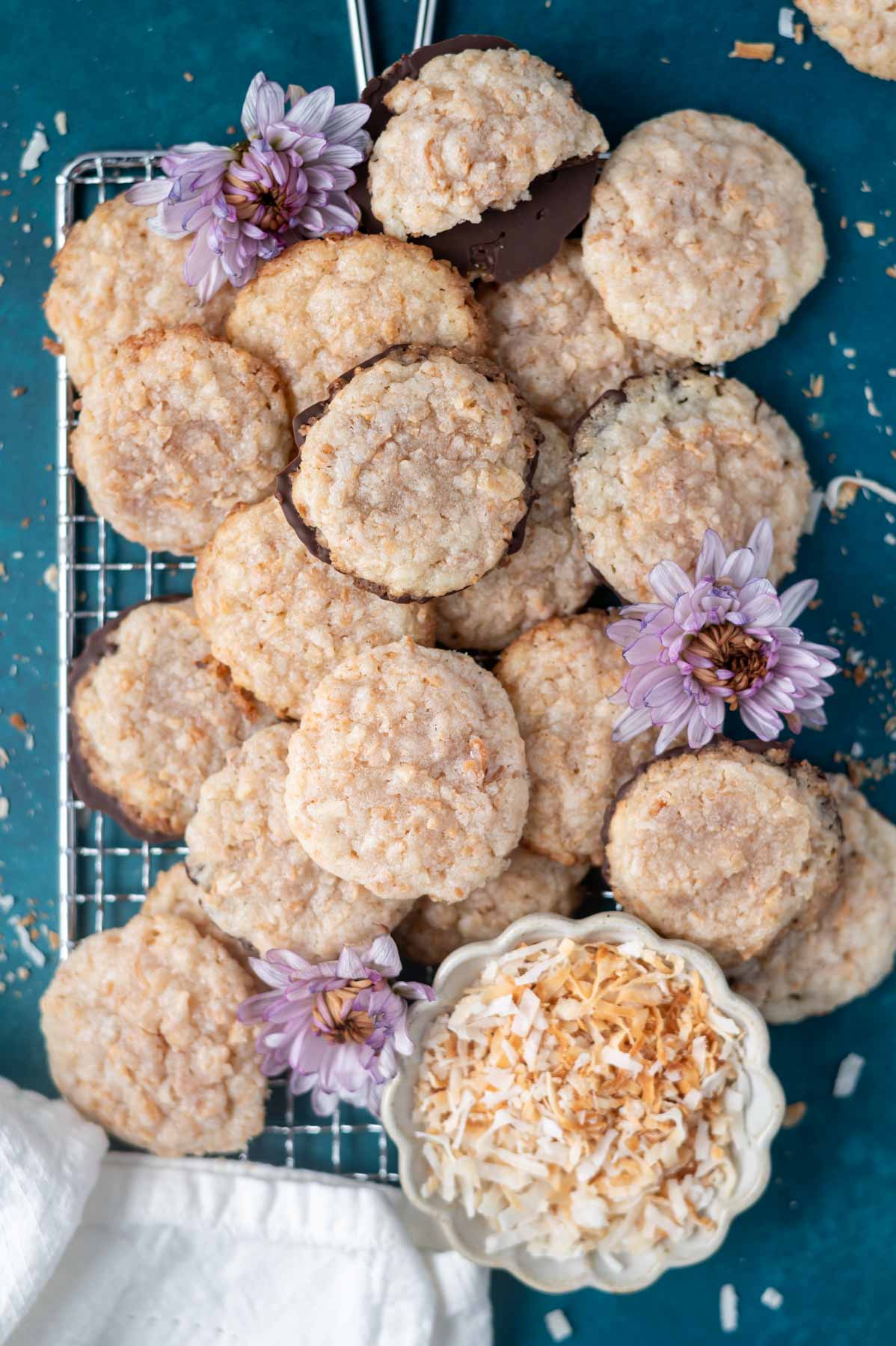 crispy coconut cookies on a cooling rack with a bowl of toasted coconut and some flowers