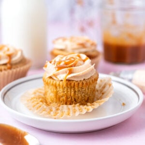 brown butter cupcake on a plate with a spoon of caramel sauce in the foreground and milk and more cupcakes in the background