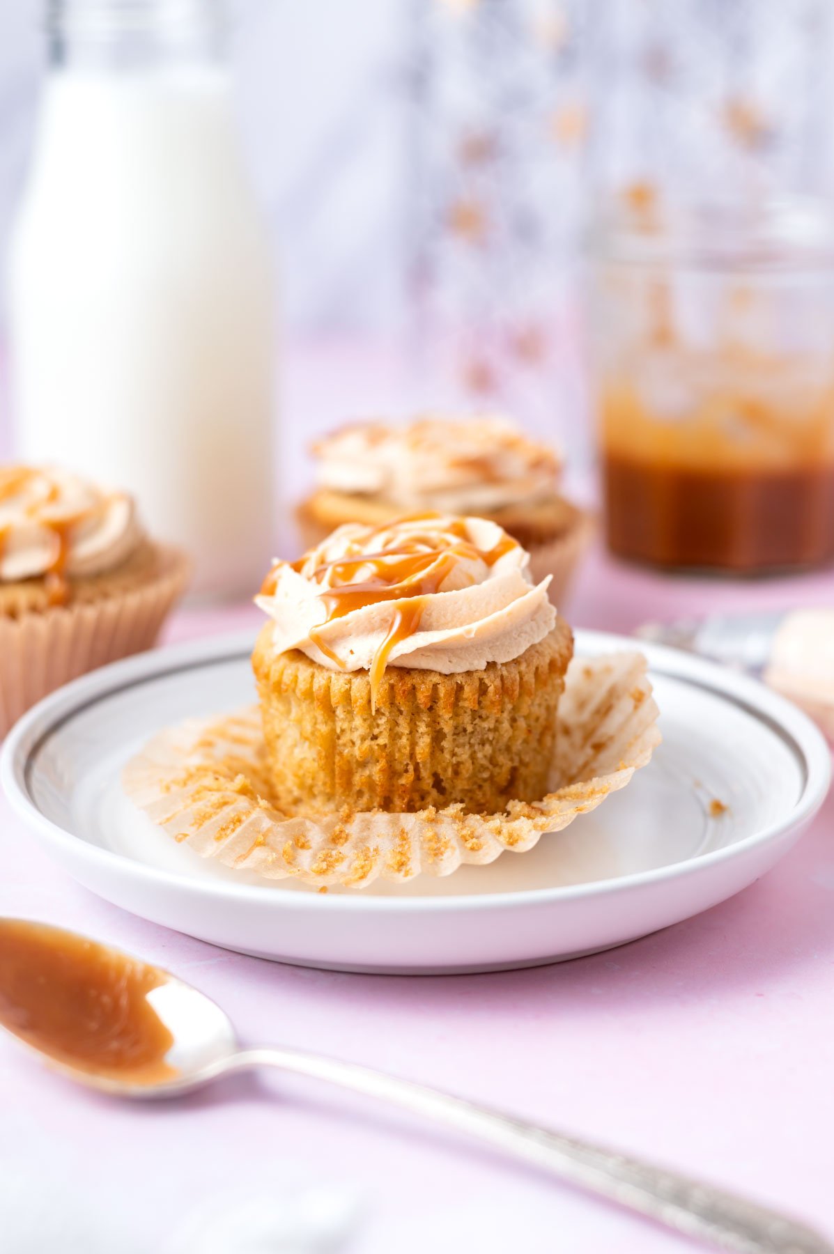 brown butter cupcake on a plate with a spoon of caramel sauce in the foreground and milk and more cupcakes in the background
