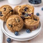 blueberry oat muffins on a plate with fresh blueberries and one muffin broken open to show soft interior