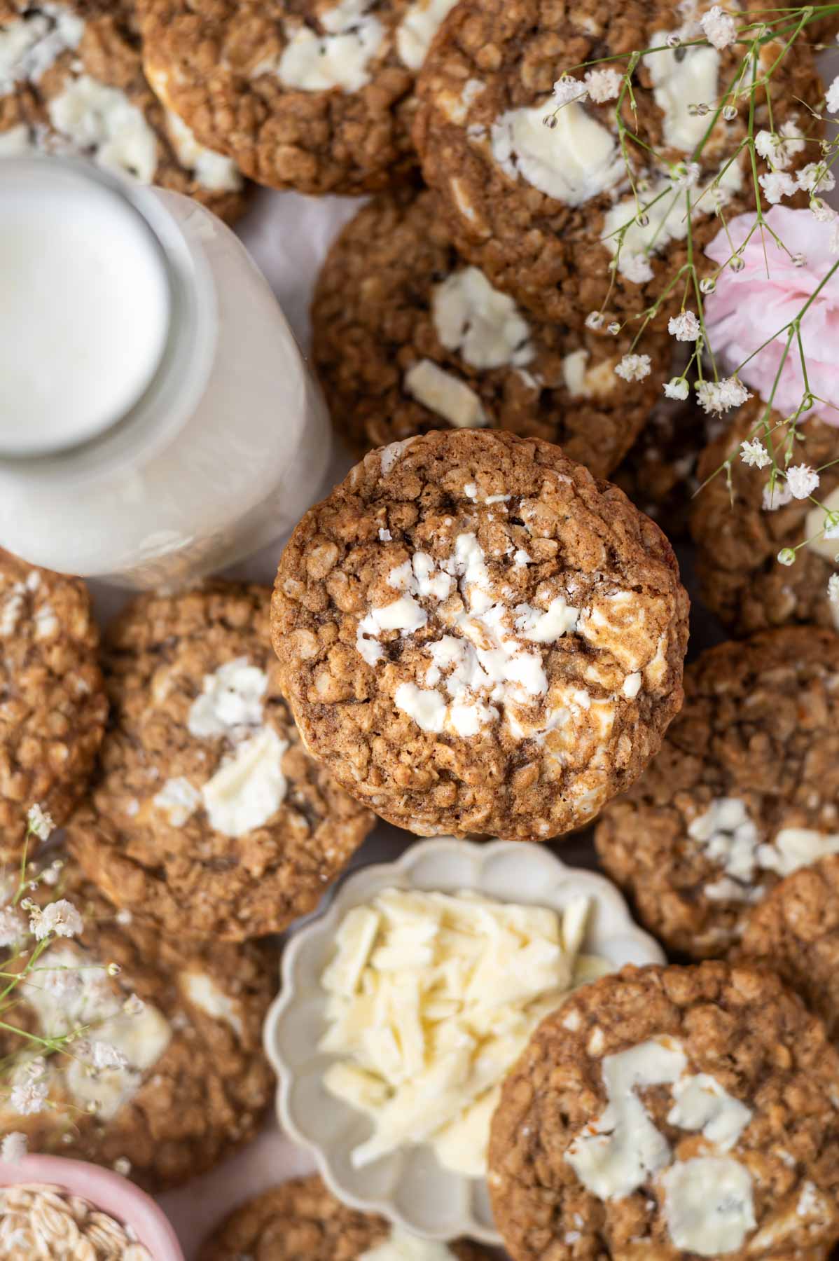 a pile of brown butter oatmeal cookies with a glass of milk and a bowl of white chocolate