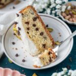 slice of cannoli cake on a plate with a fork and a bowl of pistachios and flowers in the background