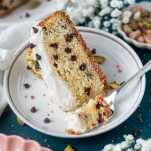 slice of cannoli cake on a plate with a fork and a bowl of pistachios and flowers in the background