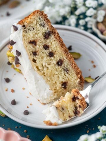 slice of cannoli cake on a plate with a fork and a bowl of pistachios and flowers in the background