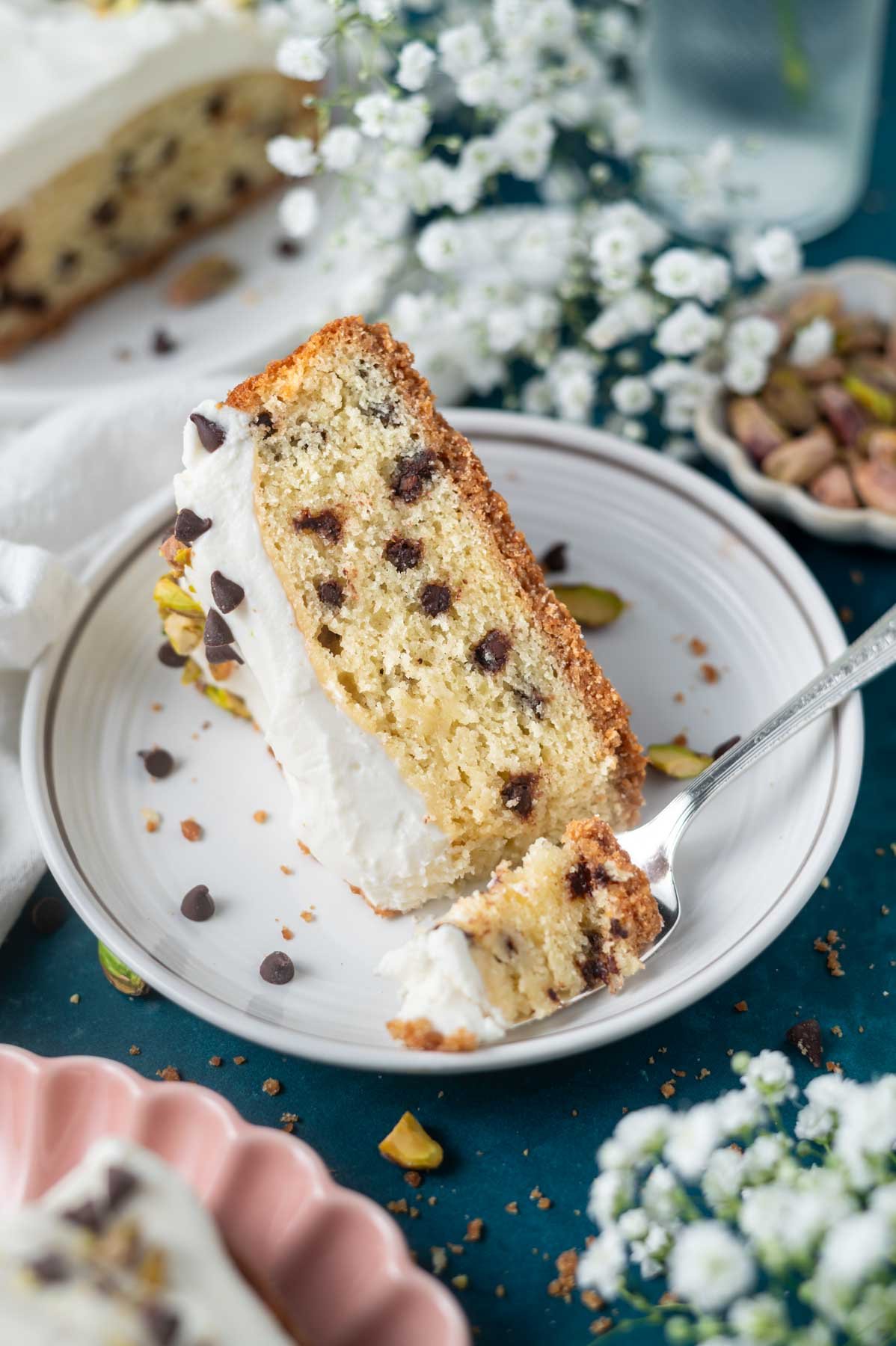 slice of cannoli cake on a plate with a fork and a bowl of pistachios and flowers in the background