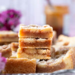 stack of shortbread caramel cookie bars on a tray with fresh dark pink flowers and caramel in the background