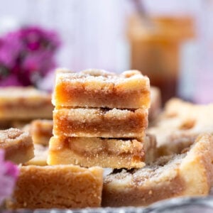 stack of shortbread caramel cookie bars on a tray with fresh dark pink flowers and caramel in the background
