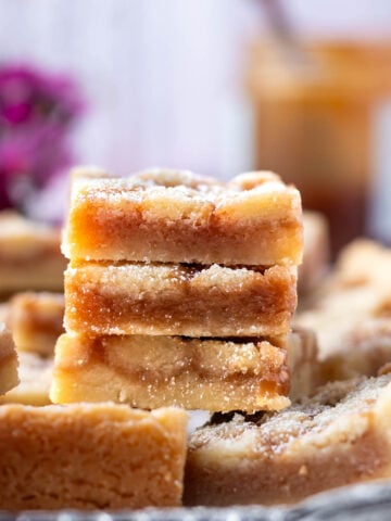 stack of shortbread caramel cookie bars on a tray with fresh dark pink flowers and caramel in the background