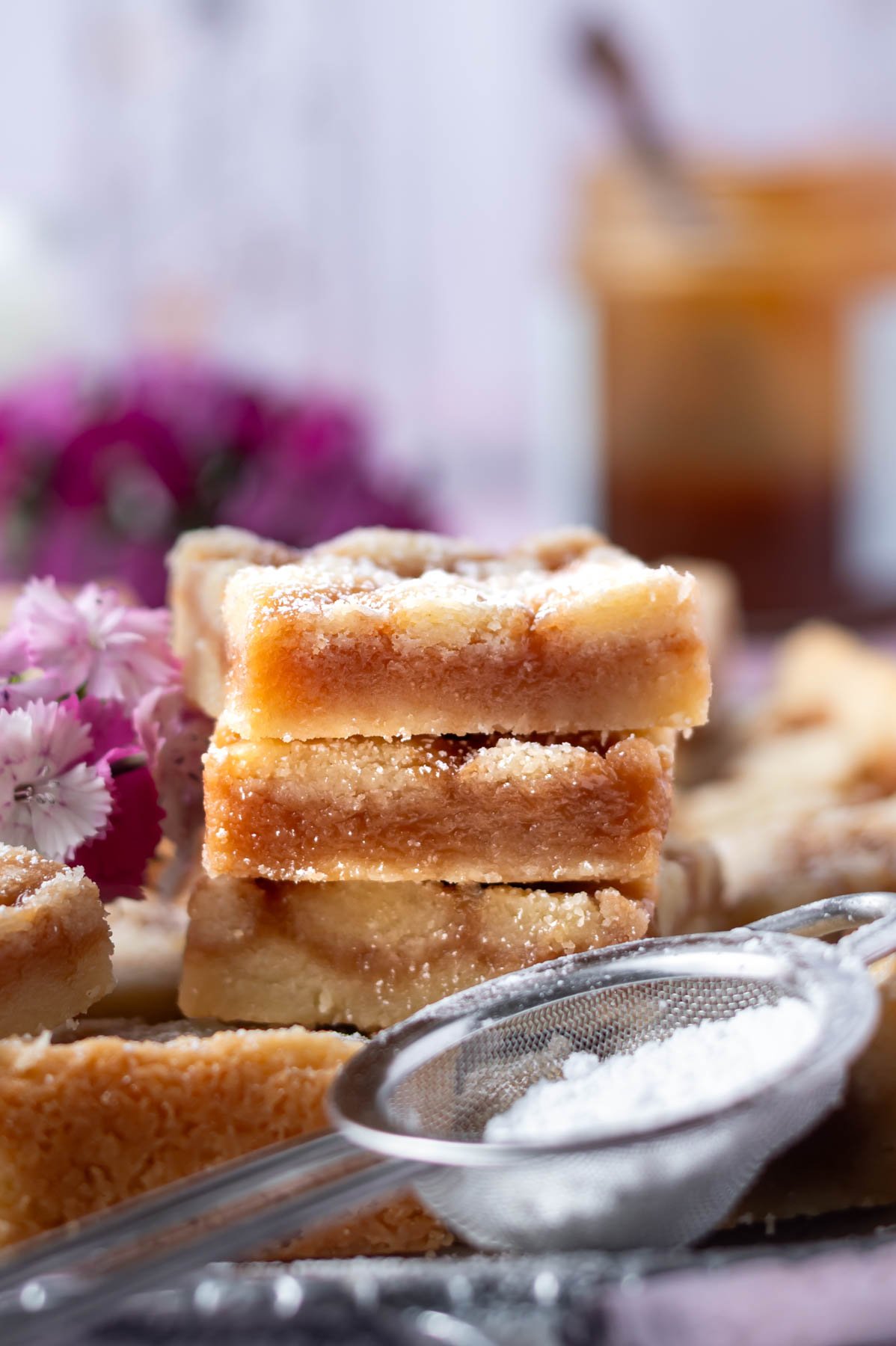 stack of cookie bars with a jar of caramel in the background and confectioners' sugar in a sifter