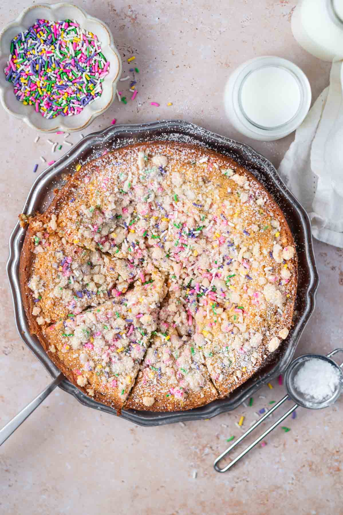 sprinkle cake on a cake platter with a bowl of sprinkles and glasses of milk