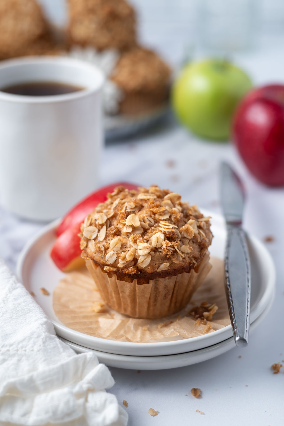 muffin on a plate with a knife with a cup of coffee and apples in the background