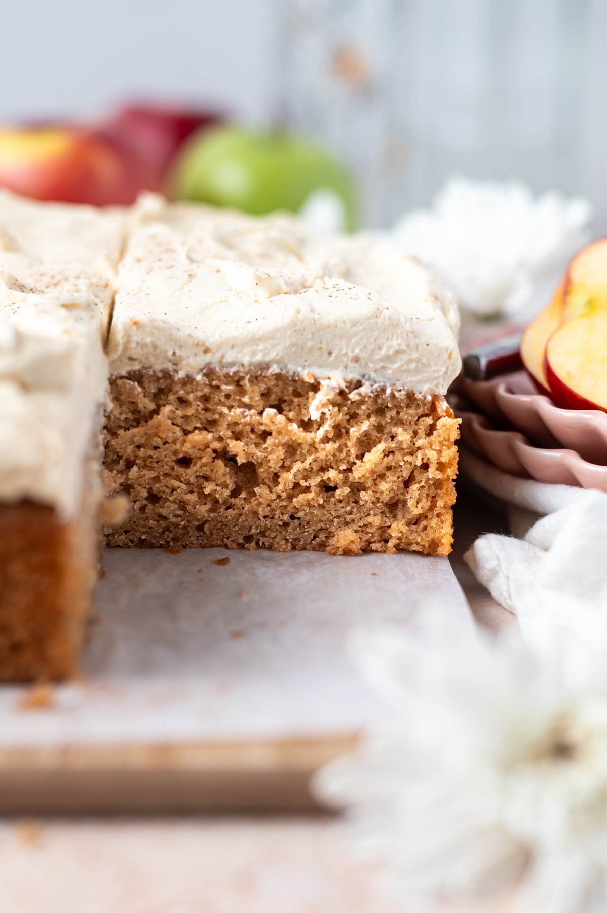 slice of cake with frosting on a parchment lined cutting board with red and green apples in the background