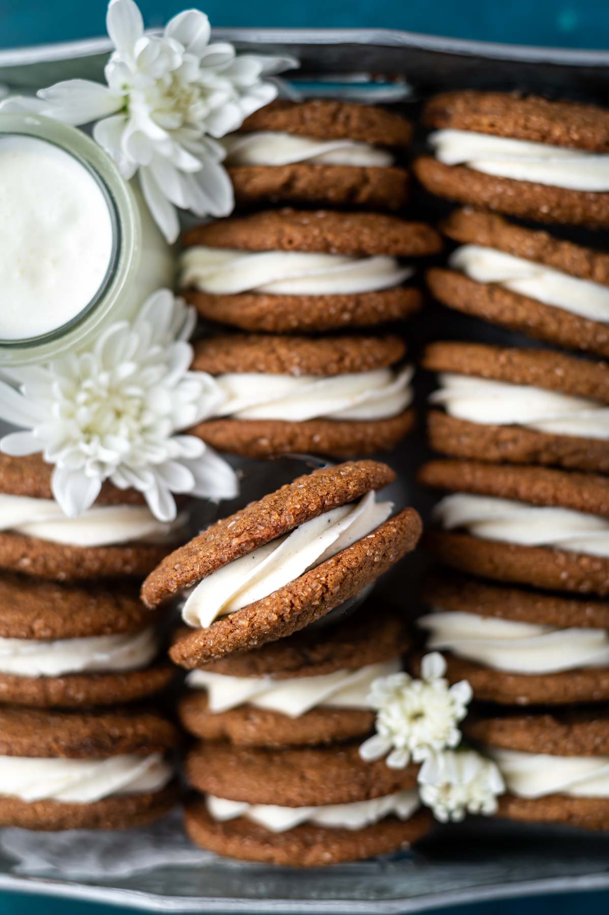 overhead look at gingerbread sandwich cookies in a tray with a glass of milk and fresh white flowers