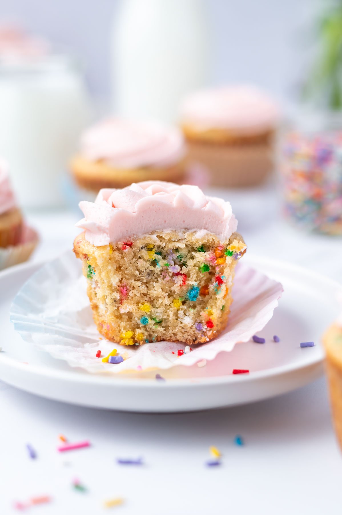 birthday cupcake with a bite missing sitting on a plate with more cupcakes and milk in the background