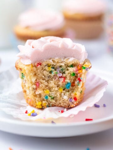 birthday cupcake with a bite missing sitting on a plate with more cupcakes and milk in the background