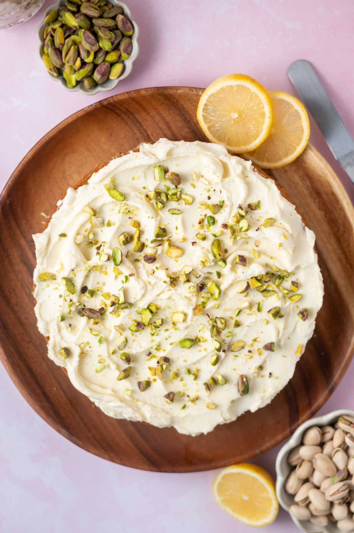 overhead look at a lemon pistachio cake on a wooden cake plate with lemon slices and bowls of pistachios