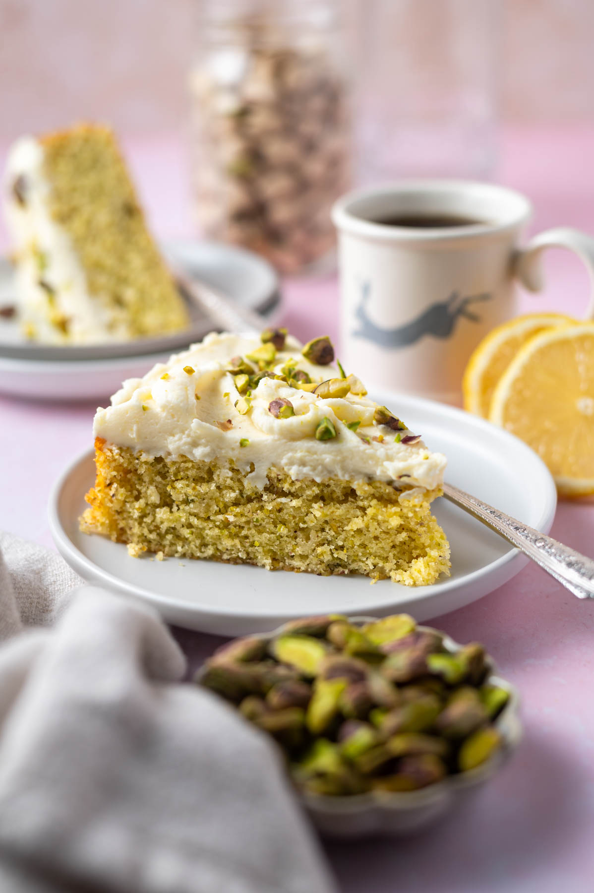slice of lemon pistachio cake on a plate with a fork and a cup of coffee, jar of pistachios and lemon slices in the background