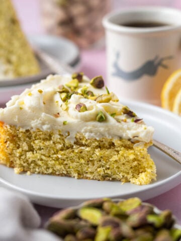 slice of lemon pistachio cake on a plate with a fork and a cup of coffee and lemon slices in the background