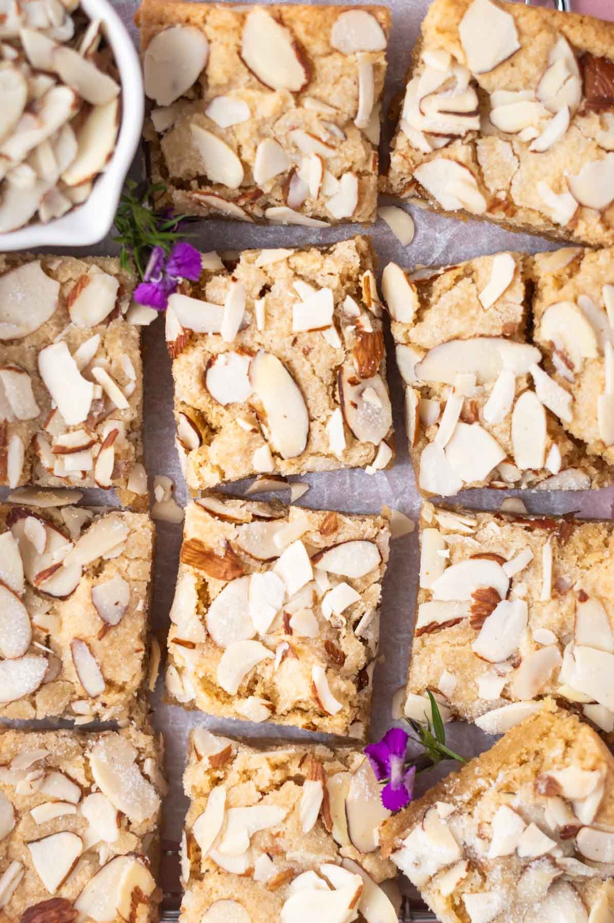 almond blondie squares on parchment paper with fresh flowers and a bowl of sliced almonds