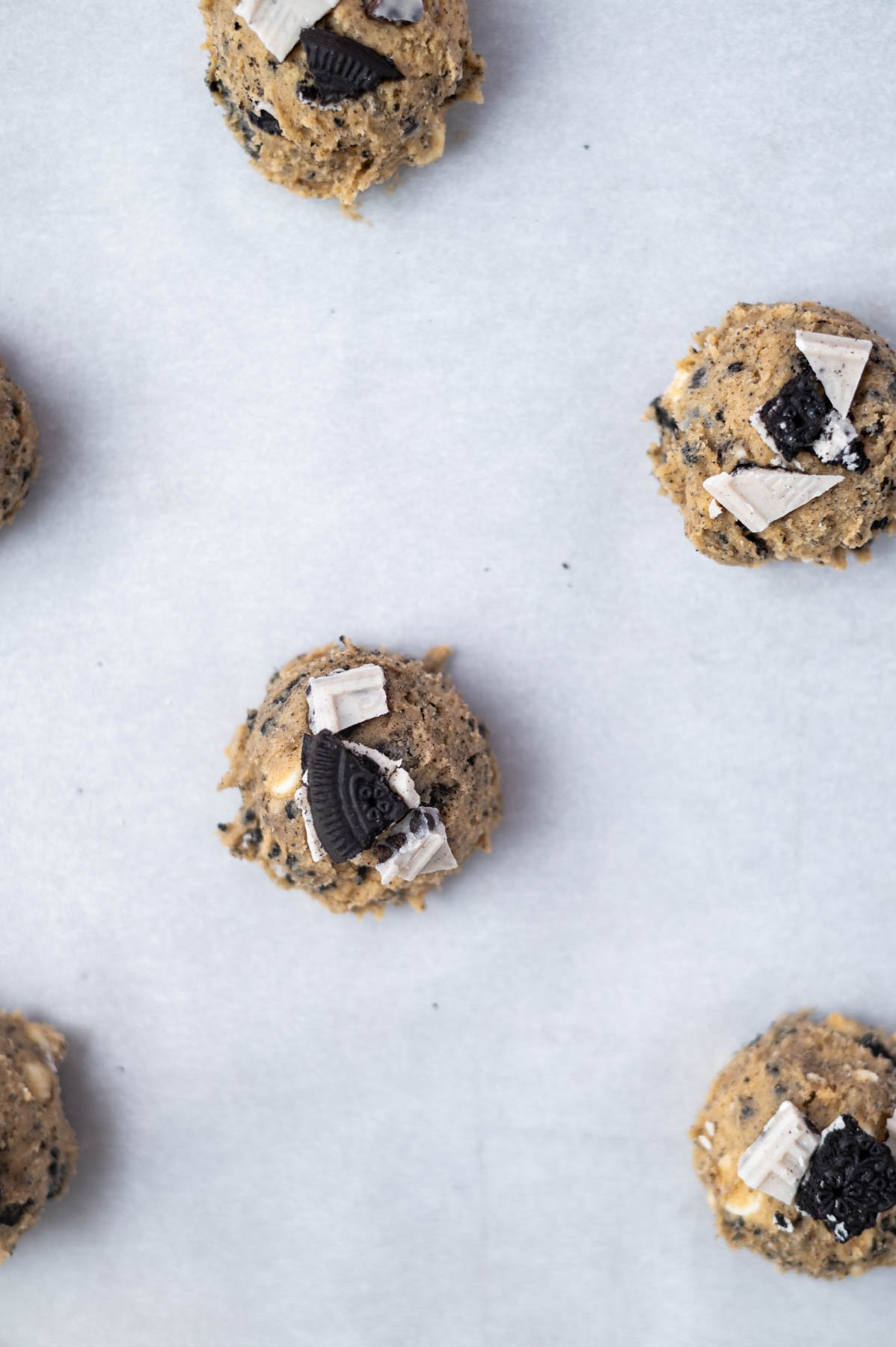 cookie dough balls on a parchment lined baking sheet with white chocolate and oreo pieces pressed into the tops