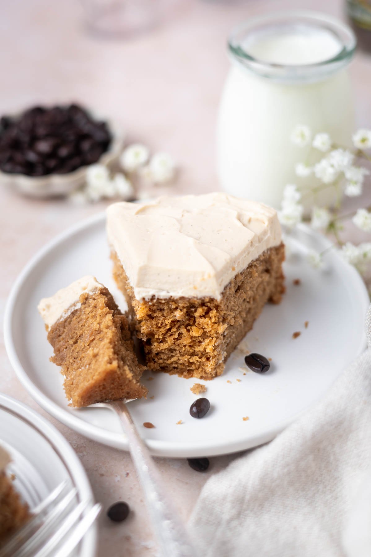 slice of cake on a plate with a bite on a fork with a glass of milk and a bowl of coffee beans in the background