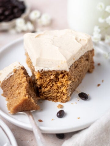 slice of cake on a plate with a bite on a fork with a glass of milk and a bowl of coffee beans in the background