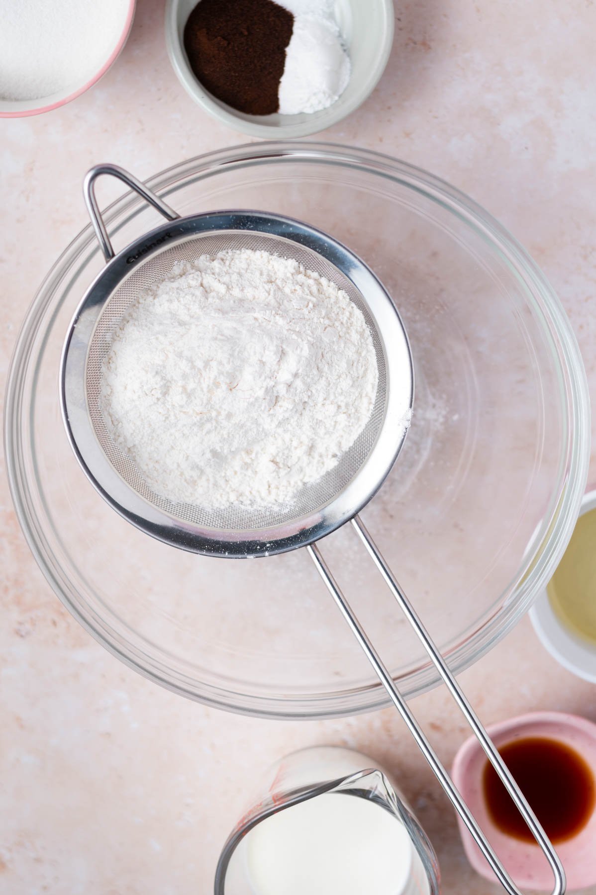 sifter with flour sitting over a glass bowl with additional ingredient surrounding