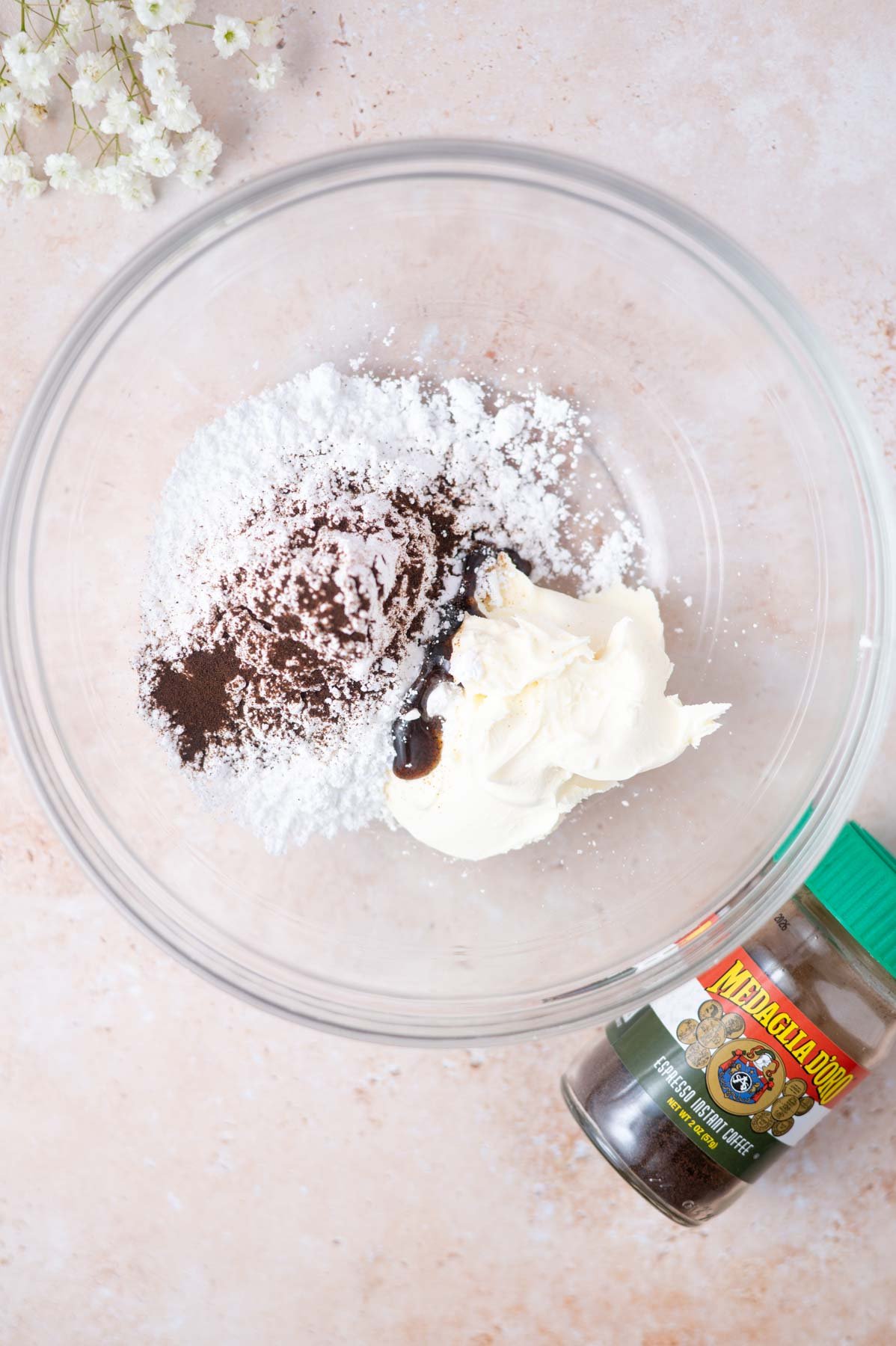 mascarpone, confectioners' sugar, vanilla bean paste and espresso powder in a glass bowl with fresh flowers and an instant espresso jar nearby