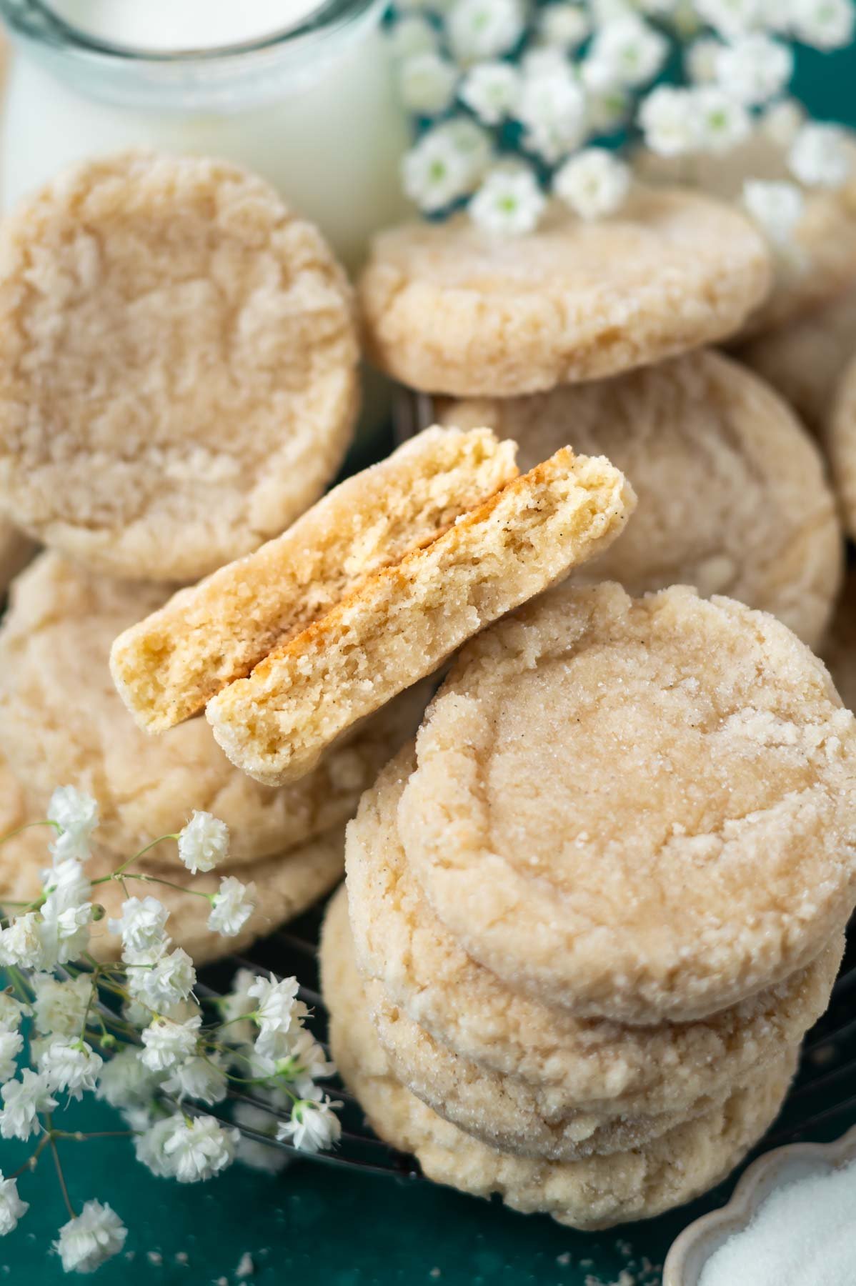 broken cookie, showing thick and soft textute, nestled among stacks of cookies with a glass of milk and fresh flowers