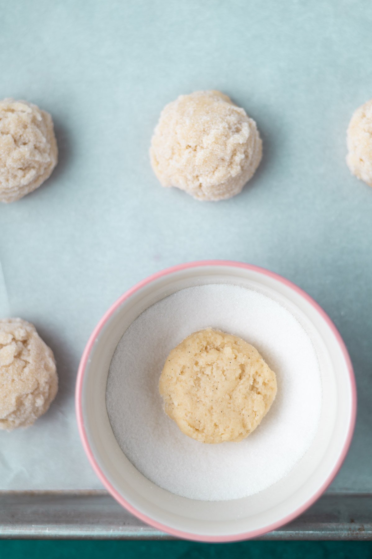 cookie dough balls on a parchment lined sheet pan with one in a bowl of sugar