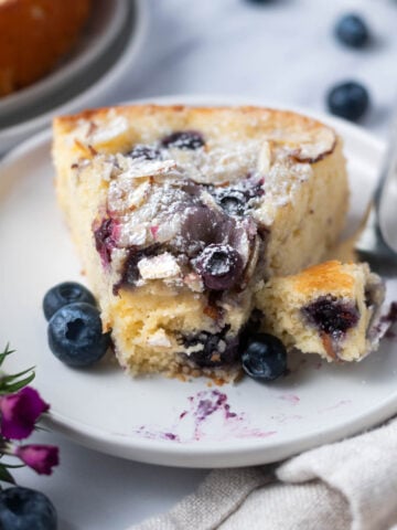 slice of blueberry almond cake on a plate with a bite on a fork and fresh blueberries