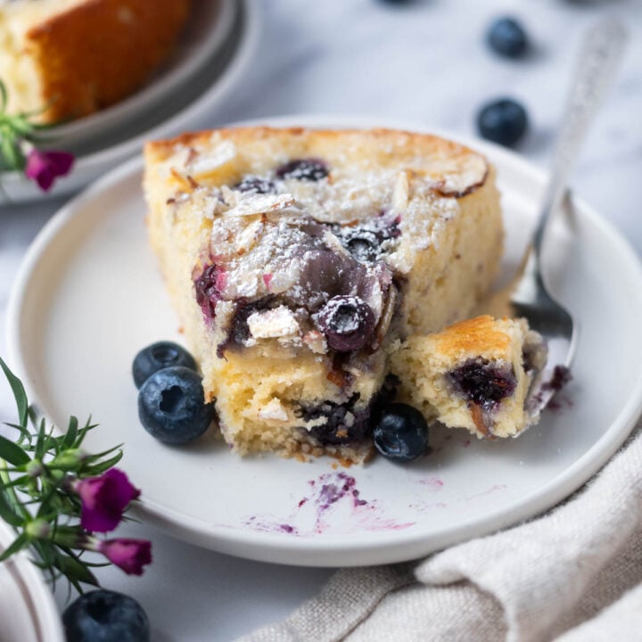 slice of blueberry almond cake on a plate with a bite on a fork and fresh blueberries
