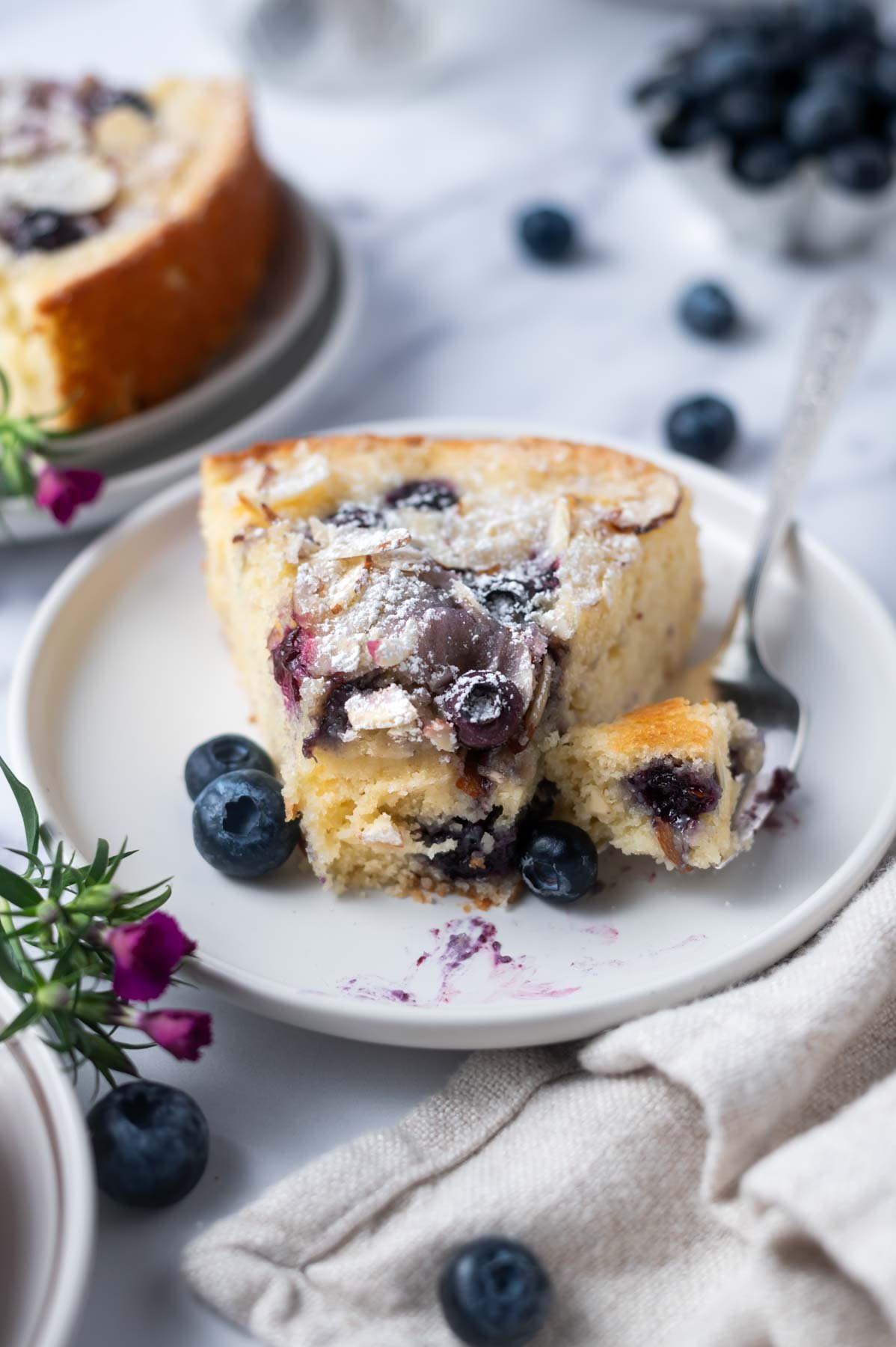 slice of blueberry almond cake on a plate with a bite on a fork and fresh blueberries