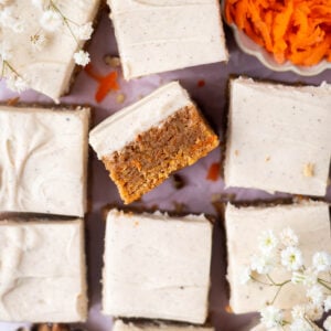 over head look at blondies with brown butter cream cheese frosting and a bowl of carrot shreds and some small white flowers