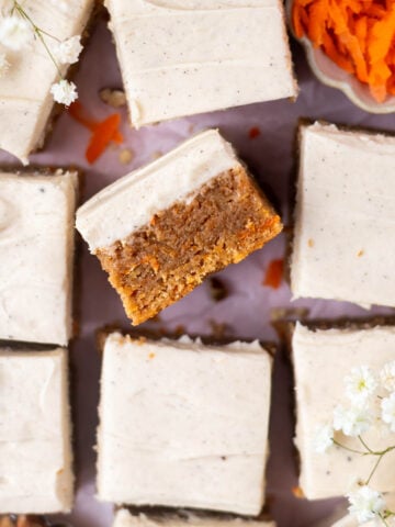 over head look at blondies with brown butter cream cheese frosting and a bowl of carrot shreds and some small white flowers