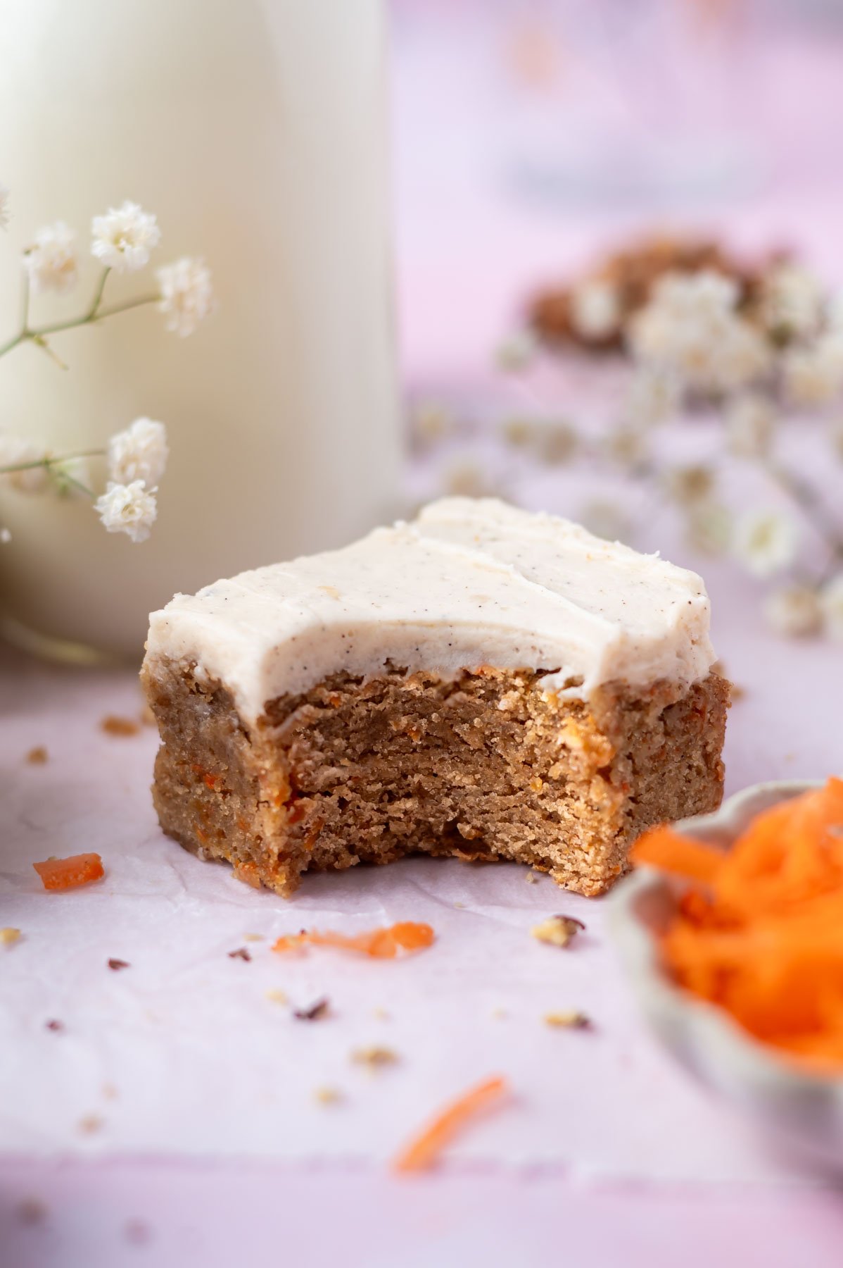 carrot cake blondie with a bite missing with a glass of milk behind and grated carrots in a bowl