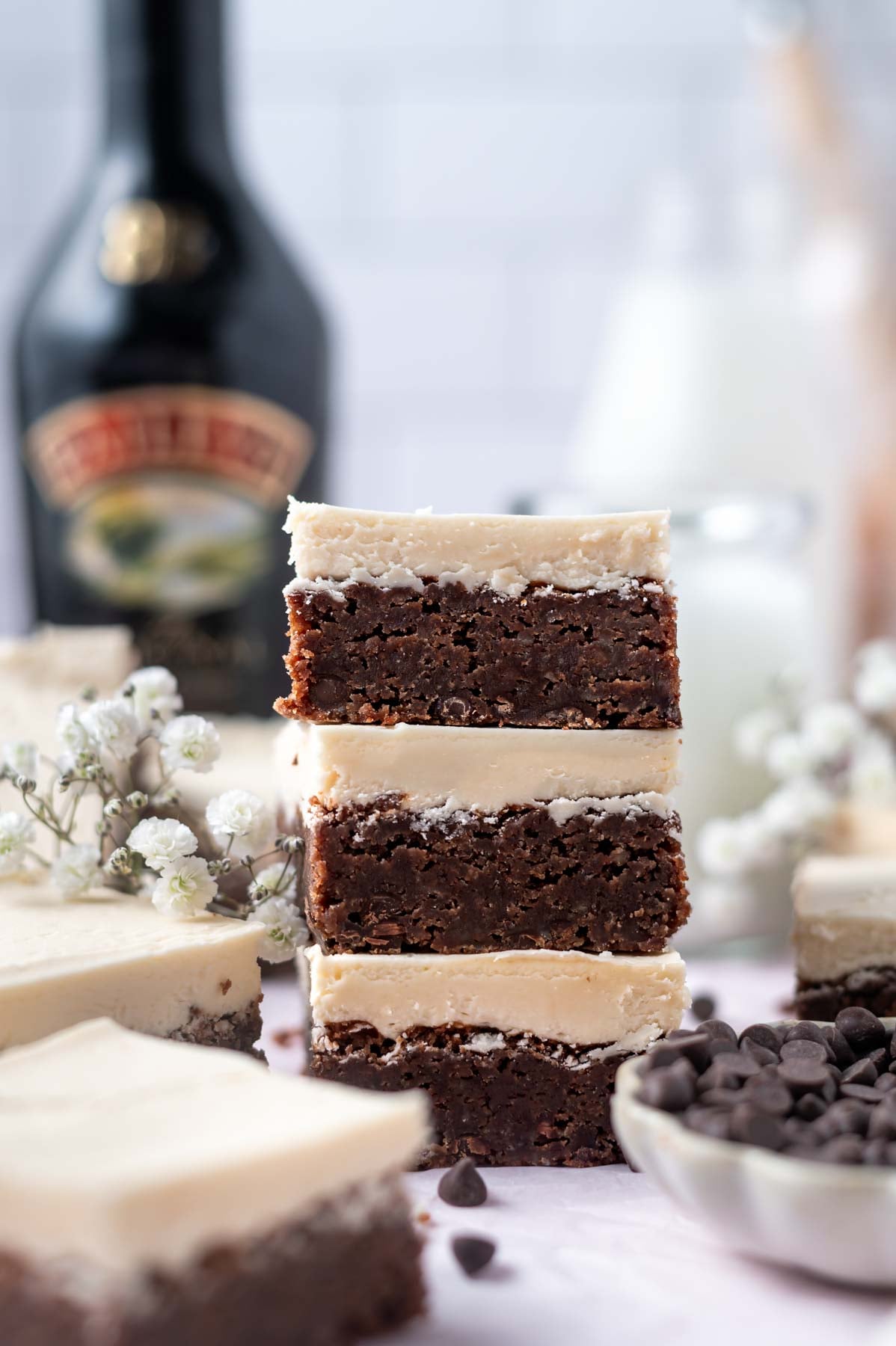 stack of frosted brownies with a bowl of chocolate chips in the foreground and milk in the background