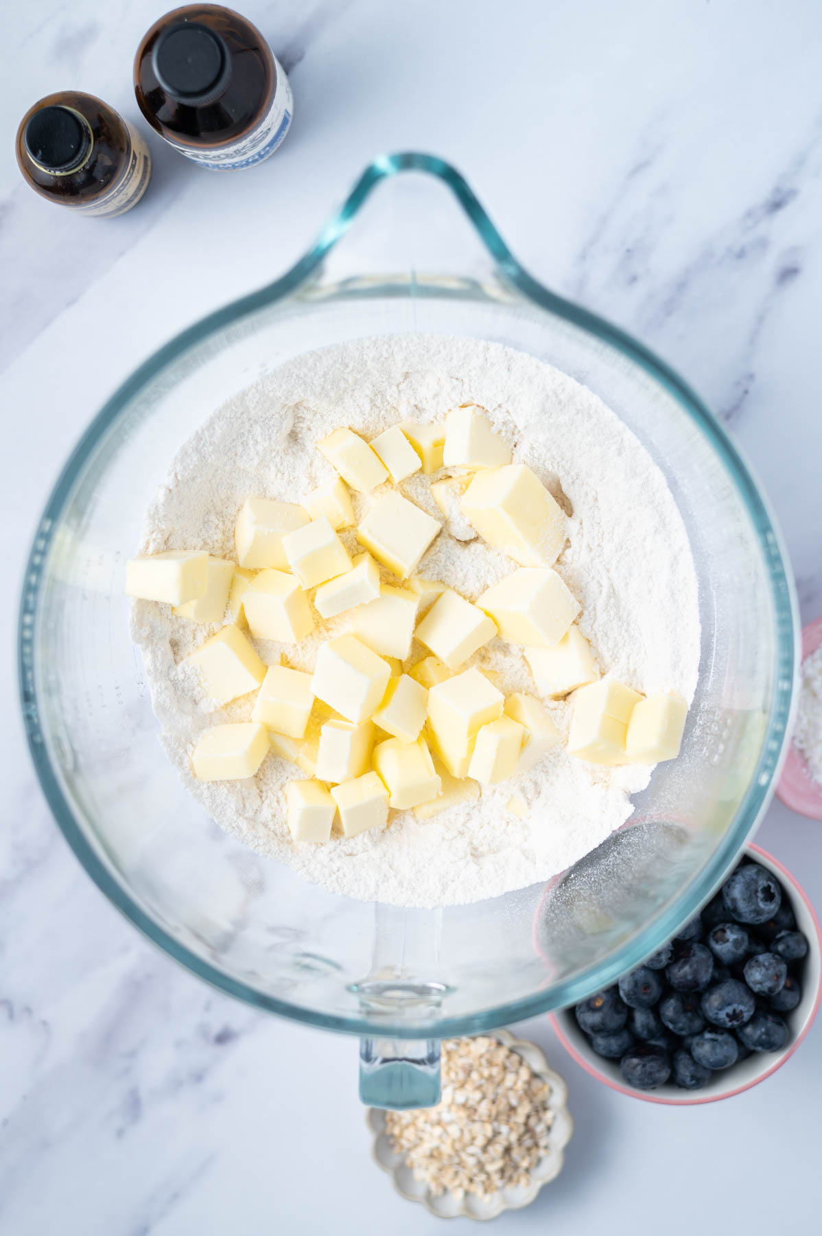 cubed butter added to flour and sugar in a mixing bowl with blueberries, oats and extracts on the side