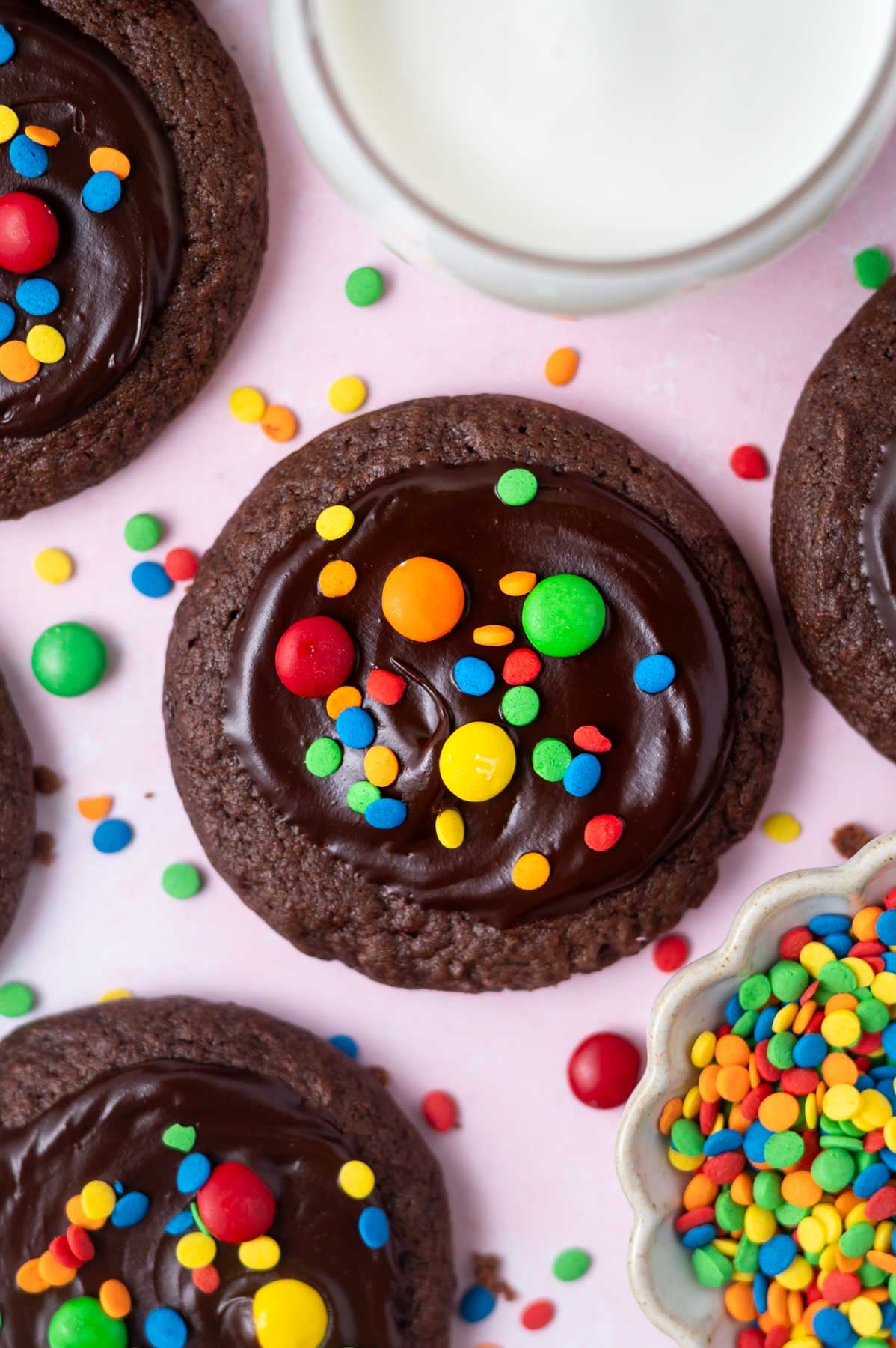 cosmic brownie cookies with ganache and colored candy topping and a glass of milk and bowl of sprinkles